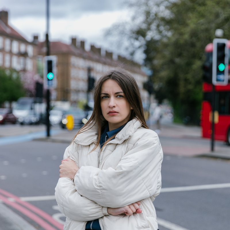 Junge Frau steht mit verschränkten Armen an einer Londoner Straße; roter Doppeldeckerbus im Hintergrund